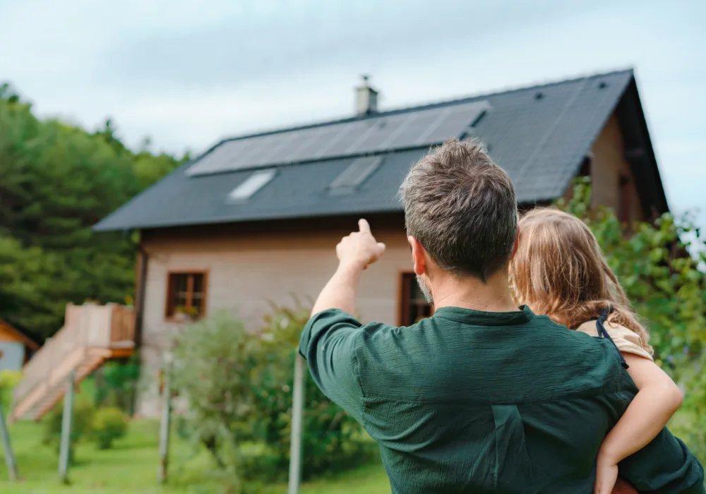 Ein Vater hält sein Kind auf dem Arm und zeigt ihm die Solarpanels auf ihrem Hausdach. Dabei handelt es sich um eine Eigenverbrauchsvariante