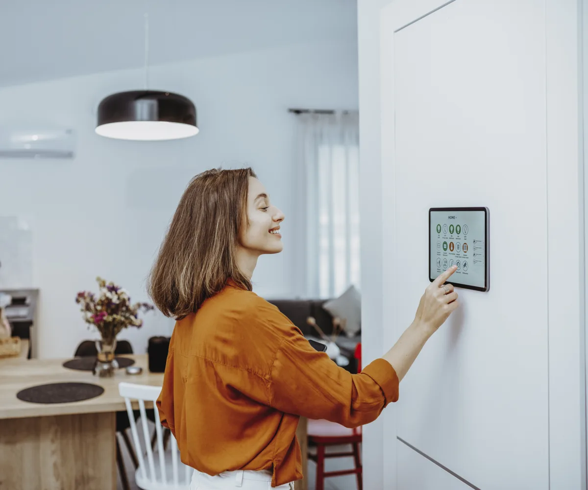 Eine junge Frau in orangenem Pullover steht im Wohnzimmer an der Wand und bedient via Touch Panel die Funktionen ihres Smarthomes.