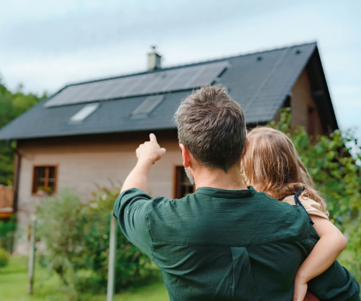 Ein Vater hält sein Kind auf dem Arm und zeigt ihm die Solarpanels auf ihrem Hausdach. Dabei handelt es sich um eine Eigenverbrauchsvariante