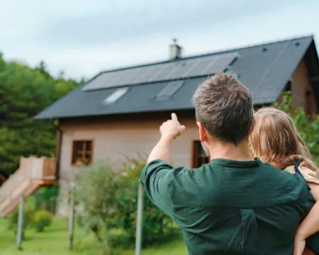 Ein Vater hält sein Kind auf dem Arm und zeigt ihm die Solarpanels auf ihrem Hausdach. Dabei handelt es sich um eine Eigenverbrauchsvariante