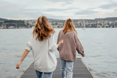 Zwei junge Frauen aus der Perspektive von hinten. Sie betreten in beschwingtem Gang einen Gittersteg, der auf den Vierwaldstättersee hinausführt. Im Hintergrund ist ein Segelboot und am Horizont das Ufer mit Teilen der Stadt Luzern zu sehen.