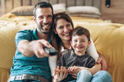 Vater, Mutter und Sohn sitzen im Wohnzimmer, der Vater richtet die Fernbedienung auf den Fernseher.
