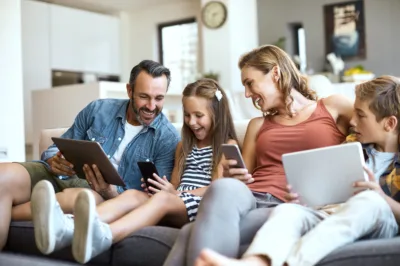 Eine Familie sitzt im Wohnzimmer auf dem Sofa. Vater, Tochter, Mutter, Sohn halten ein Tablet oder ein Smartphone in der Hand. Die Atmosphäre ist entspannt, alle lachen und sind vergnügt.