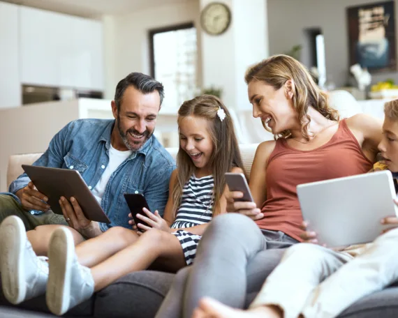 Eine Familie sitzt im Wohnzimmer auf dem Sofa. Vater, Tochter, Mutter, Sohn halten ein Tablet oder ein Smartphone in der Hand. Die Atmosphäre ist entspannt, alle lachen und sind vergnügt.