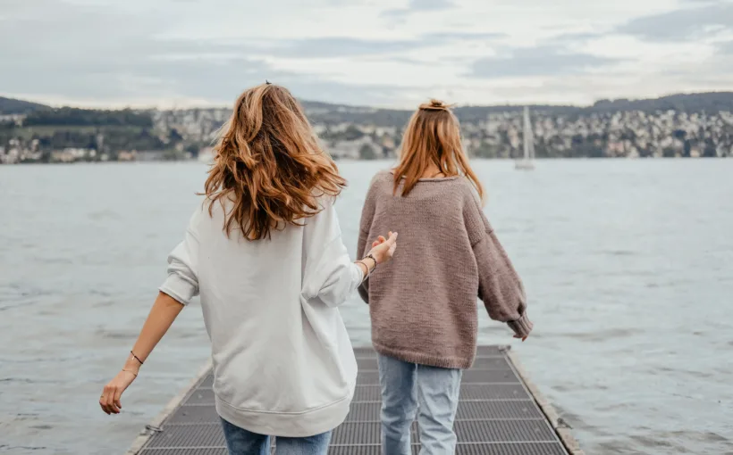 Zwei junge Frauen aus der Perspektive von hinten. Sie betreten in beschwingtem Gang einen Gittersteg, der auf den Vierwaldstättersee hinausführt. Im Hintergrund ist ein Segelboot und am Horizont das Ufer mit Teilen der Stadt Luzern zu sehen.