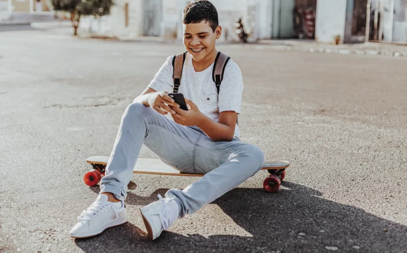 Ein Junge sitzt draussen auf der Strasse auf dem Skateboard und tippt lachend auf seinem Smartphone.