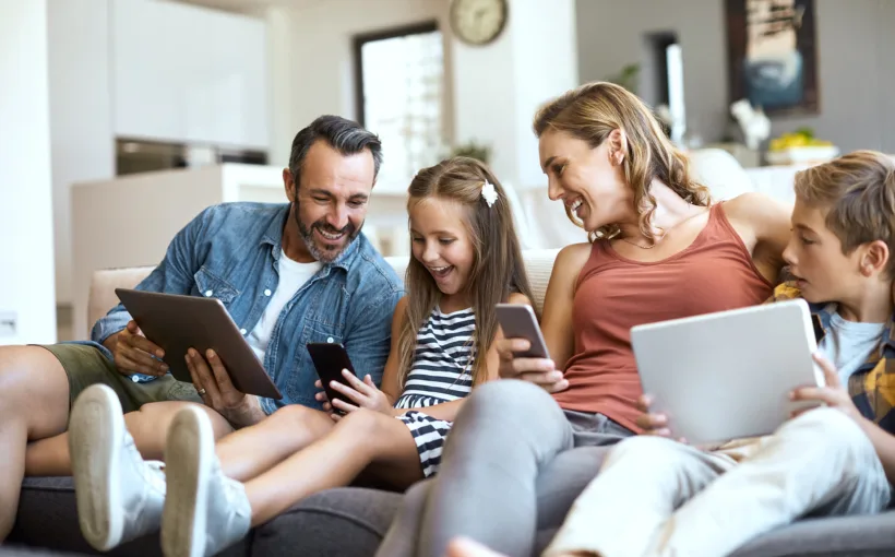 Eine Familie sitzt im Wohnzimmer auf dem Sofa. Vater, Tochter, Mutter, Sohn halten ein Tablet oder ein Smartphone in der Hand. Die Atmosphäre ist entspannt, alle lachen und sind vergnügt.