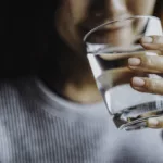 Eine Frau hält ein Glas mit Luzerner Trinkwasser in der Hand. Im Hintergrund sieht man ihr angedeutetes Lächeln. Sie führt das Glas zum Gesicht, um einen Schluck zu nehmen.