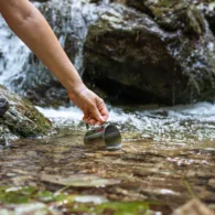 Eine Person in Wanderschuhen schöpft mit einem Trinkbecher Wasser direkt ab der Quelle eines Bergbachs