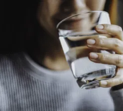 Eine Frau hält ein Glas mit Luzerner Trinkwasser in der Hand. Im Hintergrund sieht man ihr angedeutetes Lächeln. Sie führt das Glas zum Gesicht, um einen Schluck zu nehmen.